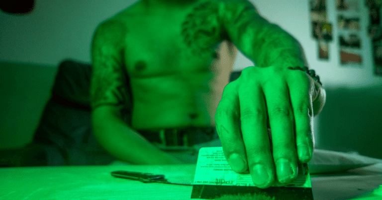 Un hombre sentado en un sofá, con la mano sobre una tarjeta, preparando lo que parece ser una dosis de droga, en un contexto de uso recreativo.