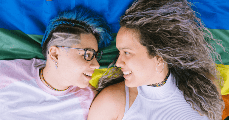 Dos chicas jóvenes tumbadas sobre una bandera arcoíris LGTBI+, mirándose y sonriendo, simbolizando el desarrollo de la identidad y la aceptación.