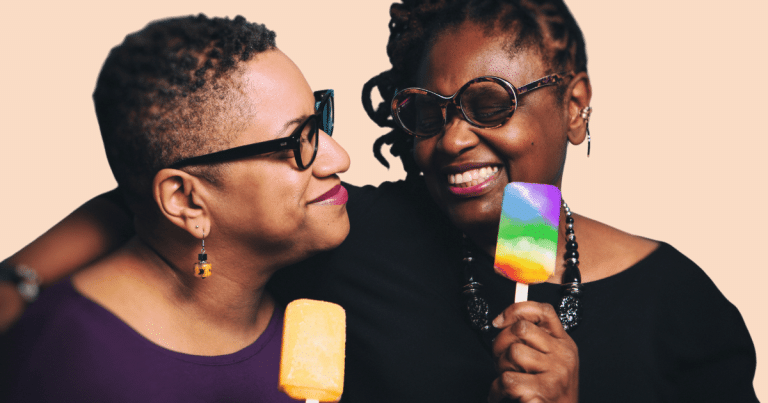 Dos mujeres en un plano cercano, una comiendo un helado con los colores del arcoíris LGTBIQ+, y ambas sonriendo y mirándose con cariño.
