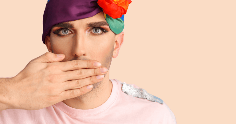 Una persona queer con maquillaje en el rostro, tapándose la boca con una mano y con un pañuelo LGTBI+ enrollado en la cabeza, mostrando una expresión de sorpresa o contención.
