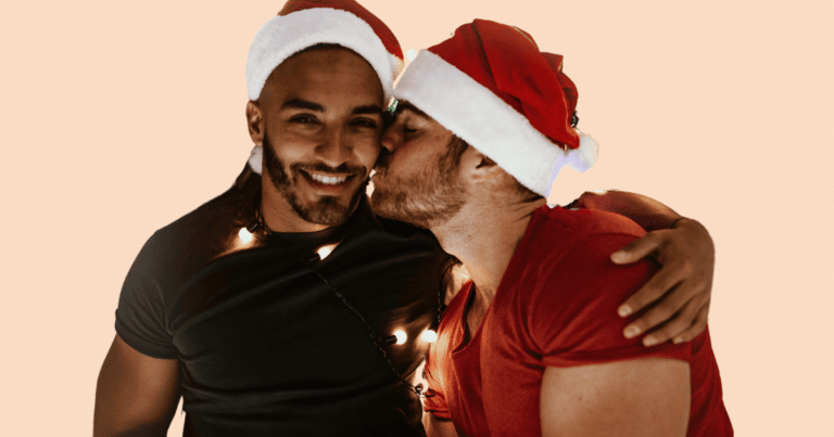 Dos hombres con gorros navideños; uno besa al otro en la mejilla mientras el otro sonríe a cámara.