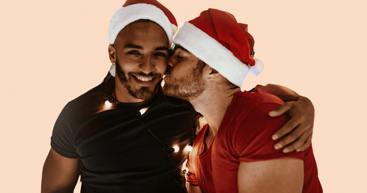 Dos hombres con gorros navideños; uno besa al otro en la mejilla mientras el otro sonríe a cámara.
