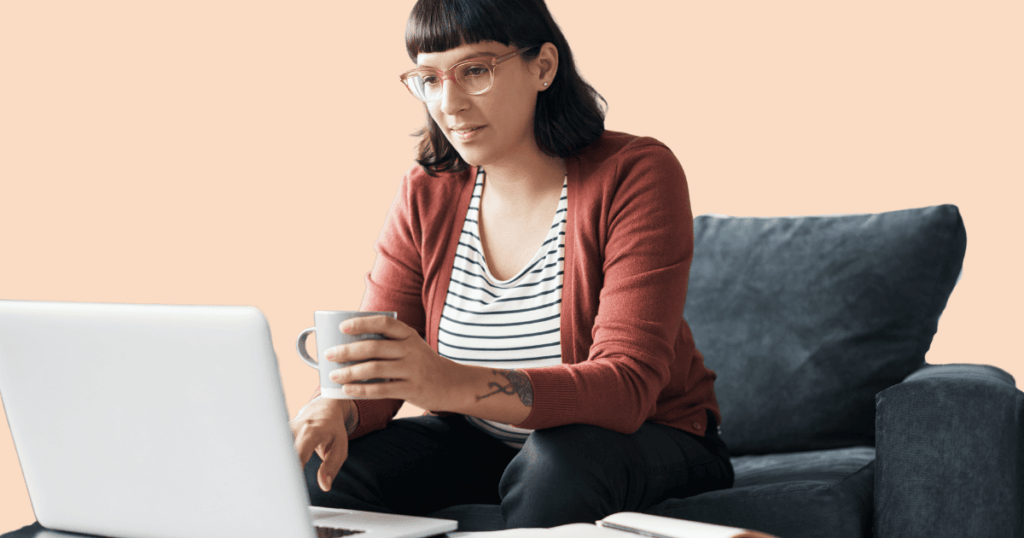 Una mujer joven sentada cómodamente en un sofá con una taza en la mano, mirando su ordenador portátil, sugiriendo una sesión de terapia online.