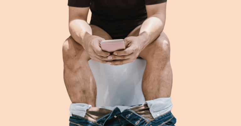 Un hombre sentado en un retrete con los pantalones bajados, mirando atentamente su teléfono móvil.