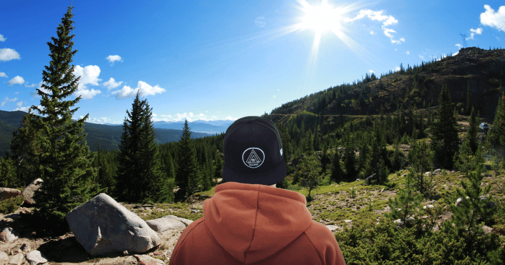 Un joven de espaldas, con gorra, observa un extenso paisaje de pinos en un día soleado, enfocado en el horizonte.