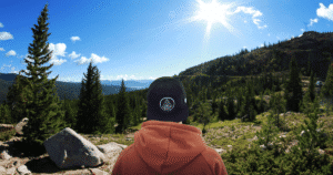 Un joven de espaldas, con gorra, observa un extenso paisaje de pinos en un día soleado, enfocado en el horizonte.