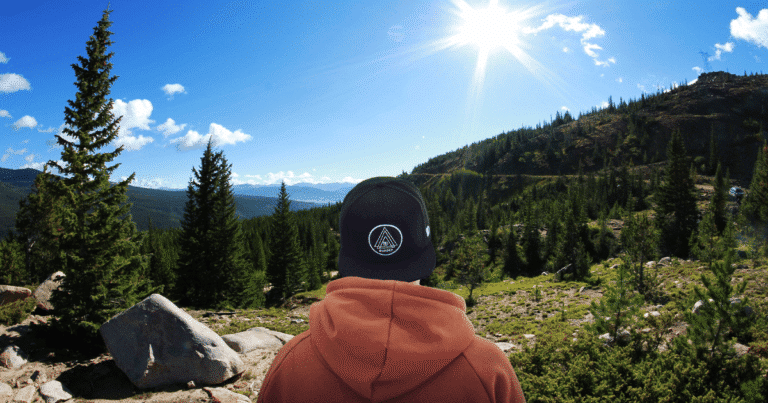 Un joven de espaldas, con gorra, observa un extenso paisaje de pinos en un día soleado, enfocado en el horizonte.