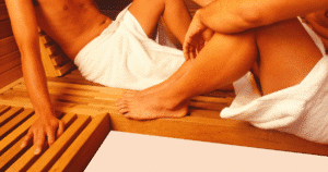 Dos hombres parcialmente visibles dentro de una sauna, cubiertos con toallas. Uno muestra su torso y el otro solo las piernas.