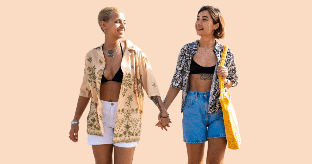 Dos mujeres lesbianas sonrientes, con ropa de verano, cogidas de la mano en un gesto de afecto y libertad, simbolizando unas vacaciones LGTBI+ seguras.